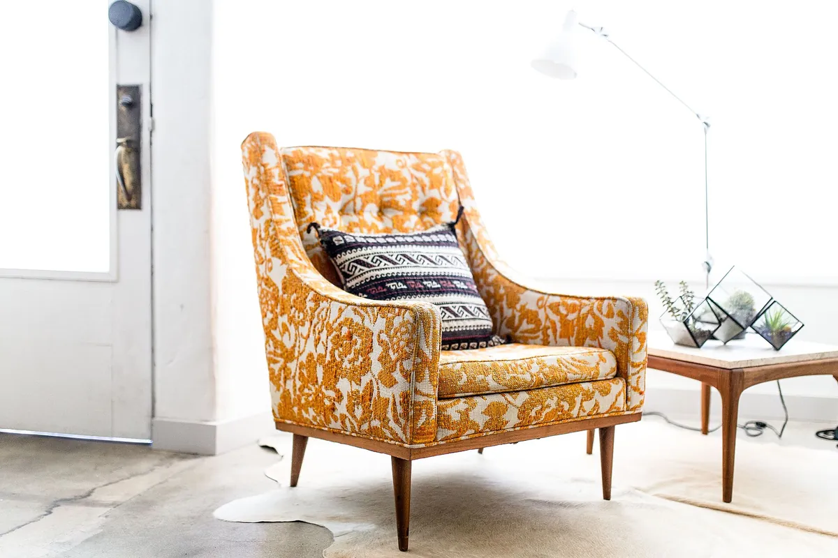 An armchair with orange floral upholstery and a striped cushion, next to a side table and a floor lamp in a bright room