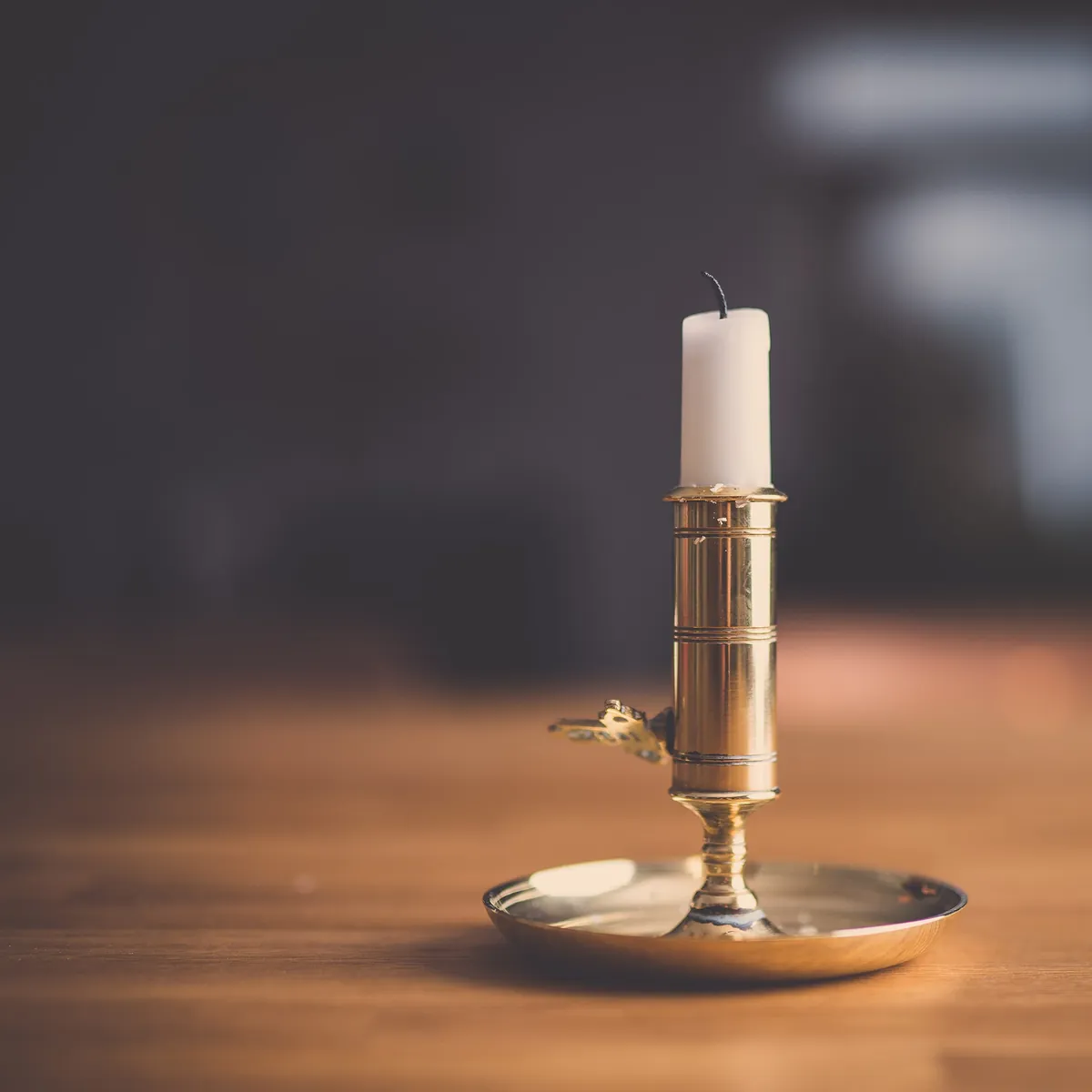 A white candle in a polished brass candle holder with a round dish, on a wooden table against a dark, blurred room