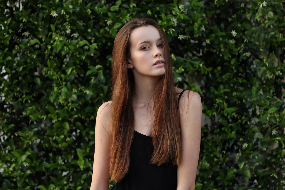 A young woman with long, straight auburn hair and a black top, in front of a hedge with small white flowers